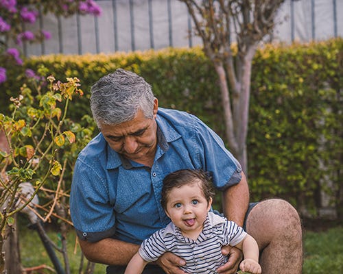 Grandfather figure holds up a toddler sticking out his tongue while trying to walk in a backyard- multigenerational homes design features allow for more family time 
