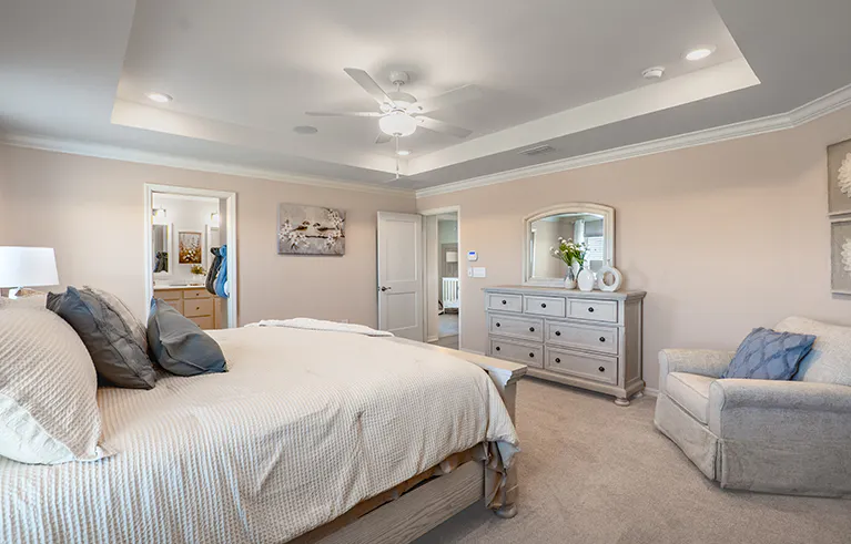 Large primary suite in the Raychel by Highland Homes, decorated in calming neutral colors, with a king bed, armchair, and large bureau.